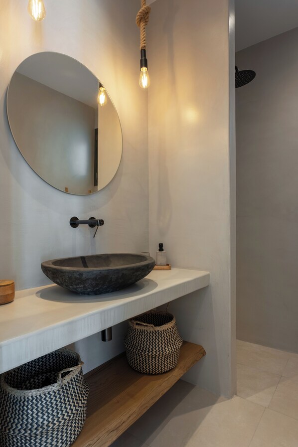 Une salle de bain moderne avec un miroir rond, un lavabo sur colonne et deux paniers en osier posés sur une étagère en bois.