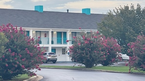 Um motel de dois andares com telhado cinza e detalhes verde-água, rodeado por arbustos floridos.