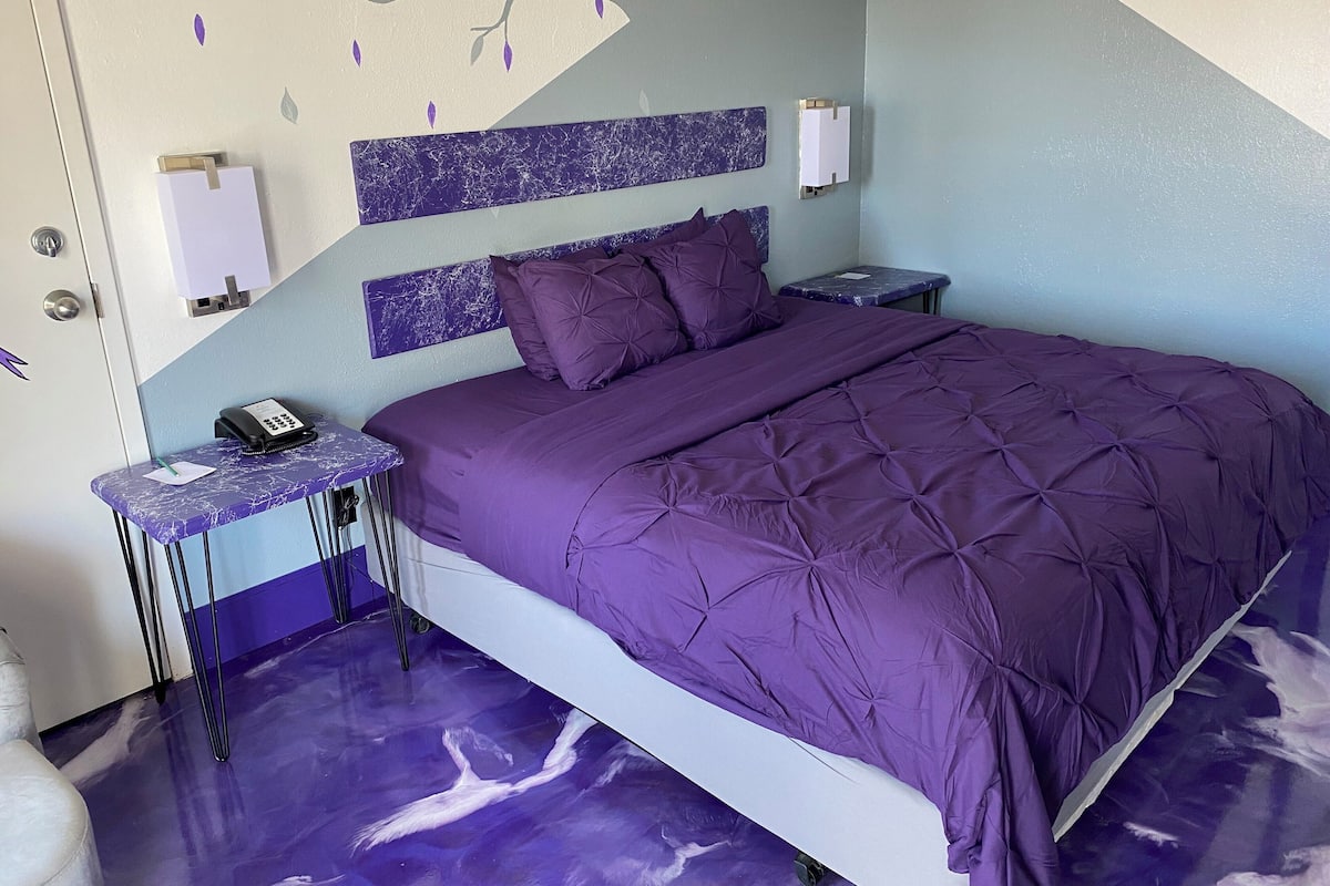 Un dormitorio con colcha morada, cabecero de mármol, mesita de noche con teléfono y una lámpara de pared.