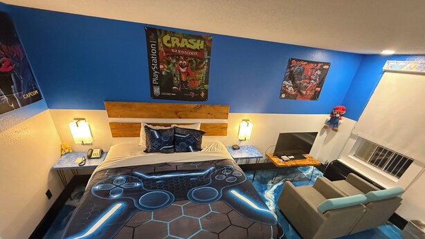 Quarto com uma cama apresentando um design de controle de videogame luminoso, um laptop em uma mesinha de cabeceira, pôsteres nas paredes e uma pequena janela.