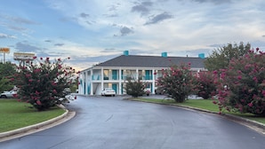 Um motel de dois andares com entrada central, estacionamento e vegetação ao redor.
