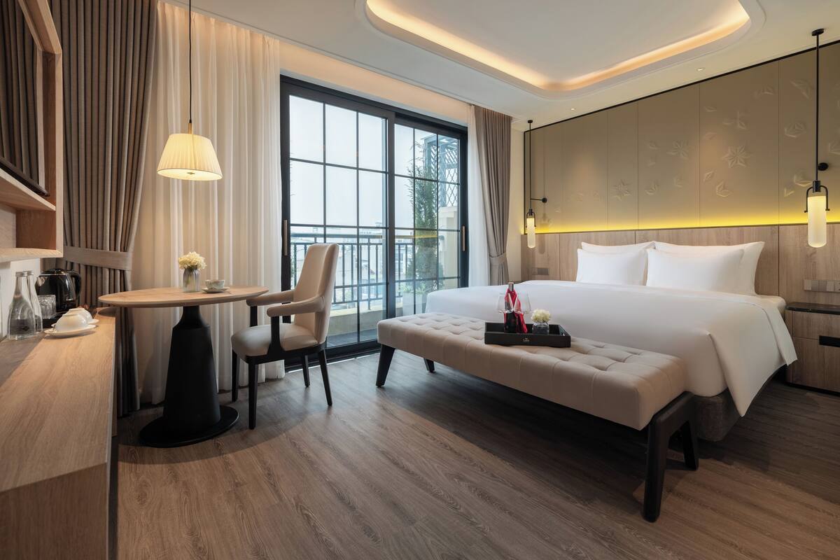 A modern hotel room with a large bed, a desk with a chair, a small table with a lamp, and a view of the city through large windows.
