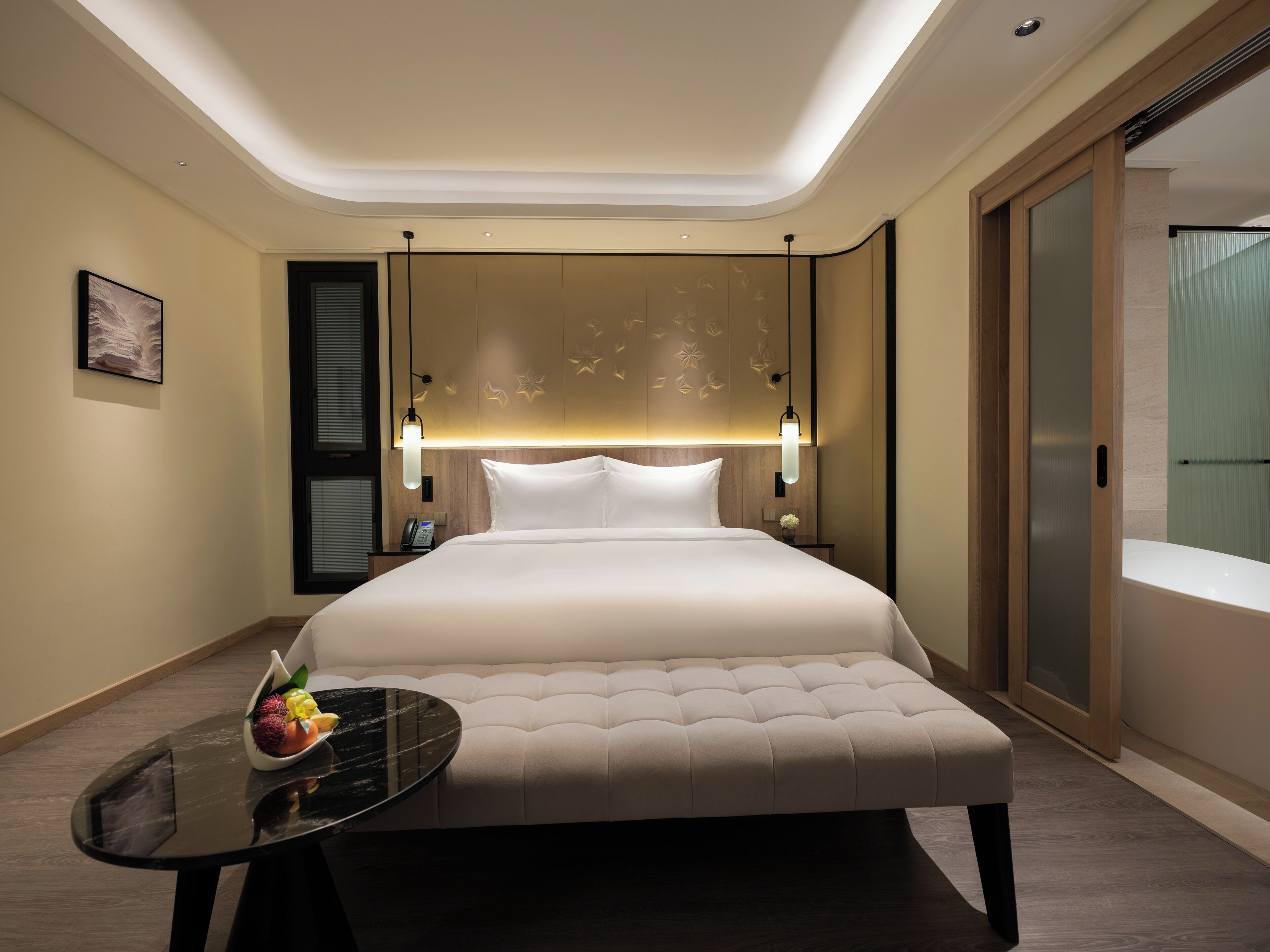 A modern hotel room with a large bed, a bedside table with a vase of flowers, and a view of the city through the window.