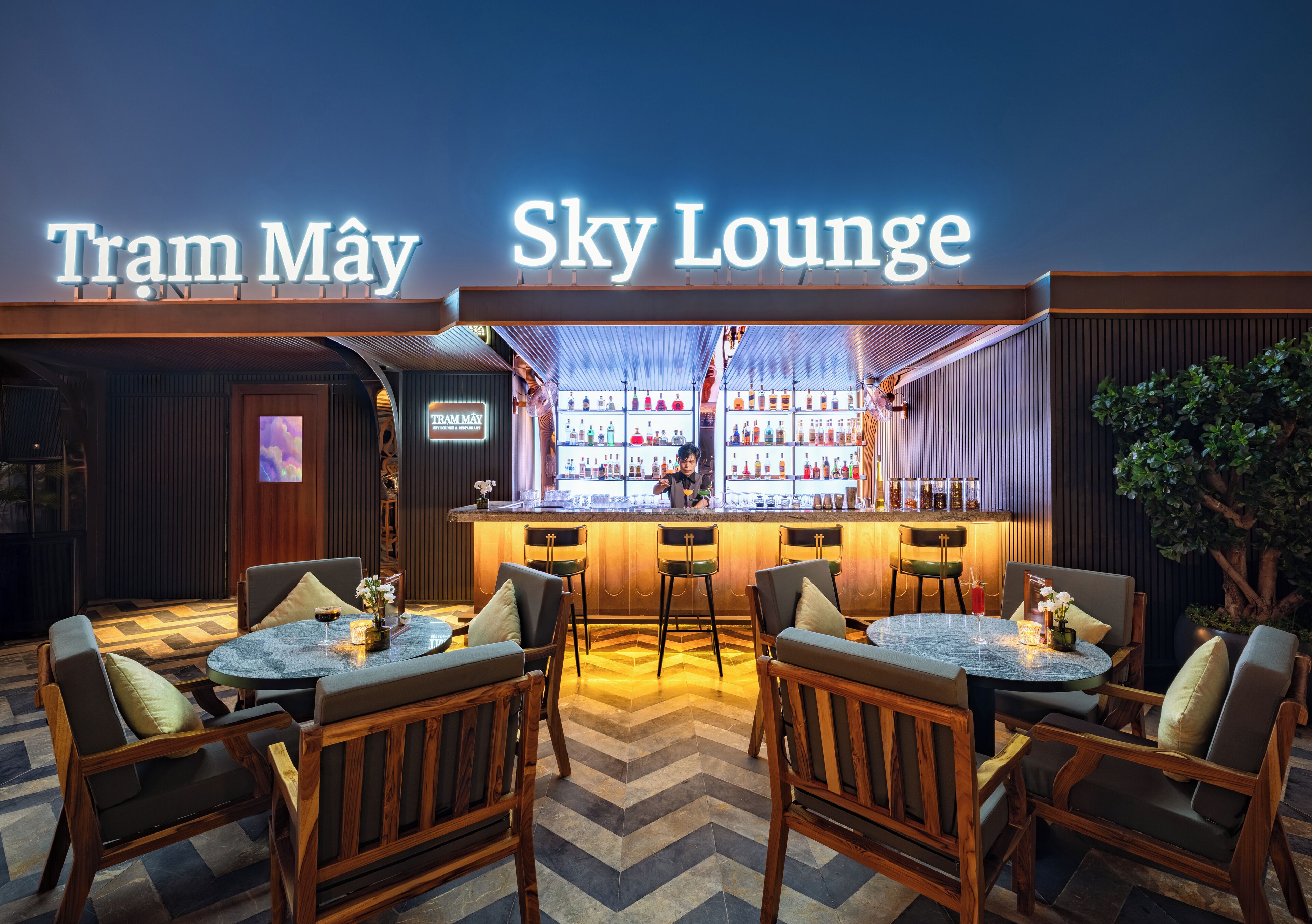 Zona de bar con un letrero que dice "Tram May Sky Lounge", con mobiliario de madera y una barra repleta de botellas.