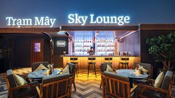 Zona de bar con un letrero que dice "Tram May Sky Lounge", con mobiliario de madera y una barra repleta de botellas.