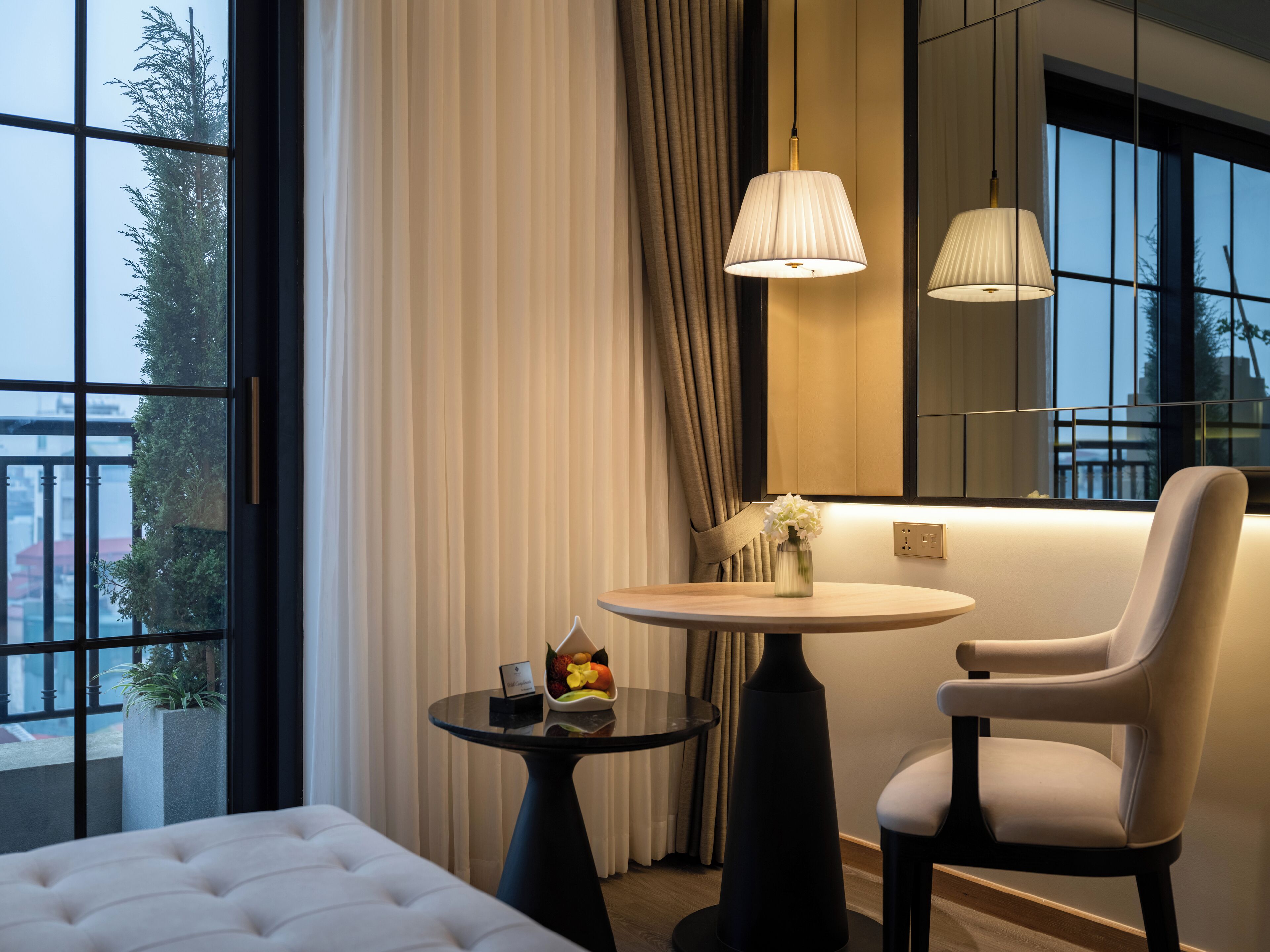 A modern hotel room with a large window, a round table with a vase of flowers, a chair, and a bed with a tufted headboard.