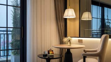 A modern hotel room with a large window, a round table with a vase of flowers, a chair, and a bed with a tufted headboard.