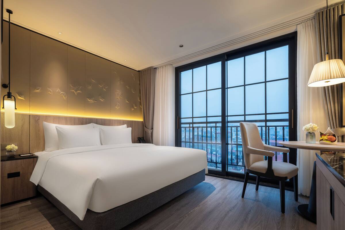 A modern hotel room with a large bed, a desk, and a view of the city through large windows.
