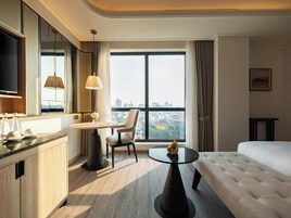 A hotel room with a bed, a desk, a chair, a TV, and a large window with a city view.