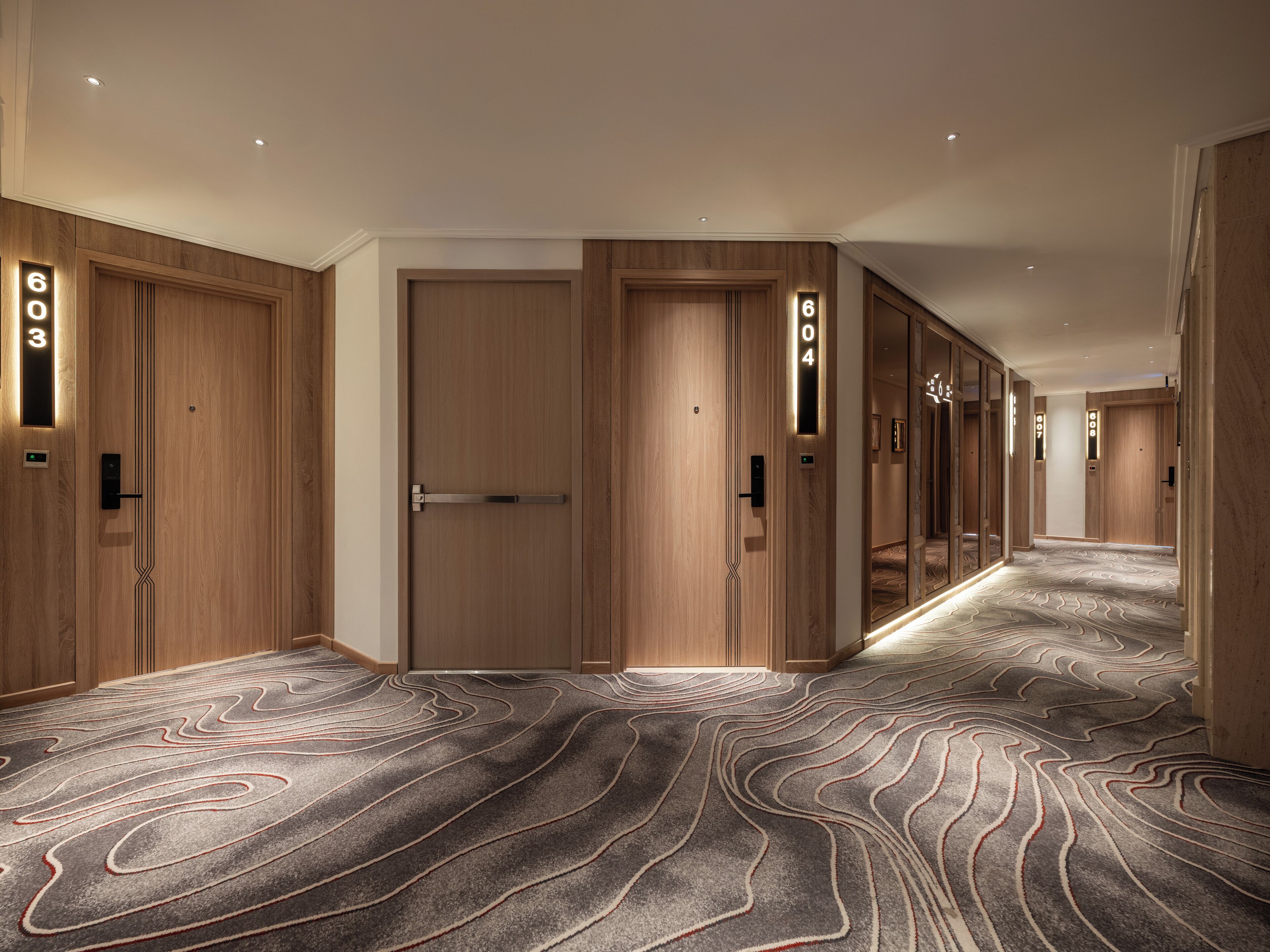 Ein Hotelkorridor mit Holztüren, beschrifteten Zimmern und einem gemusterten Teppich.