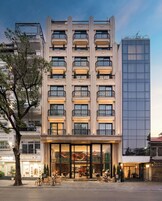 A multi-story hotel with a central entrance, balconies, and a modern glass building to the right.