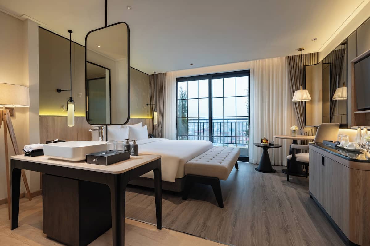 A modern hotel room with a large bed, a desk with a sink, a mirror, and a view of the city through large windows.
