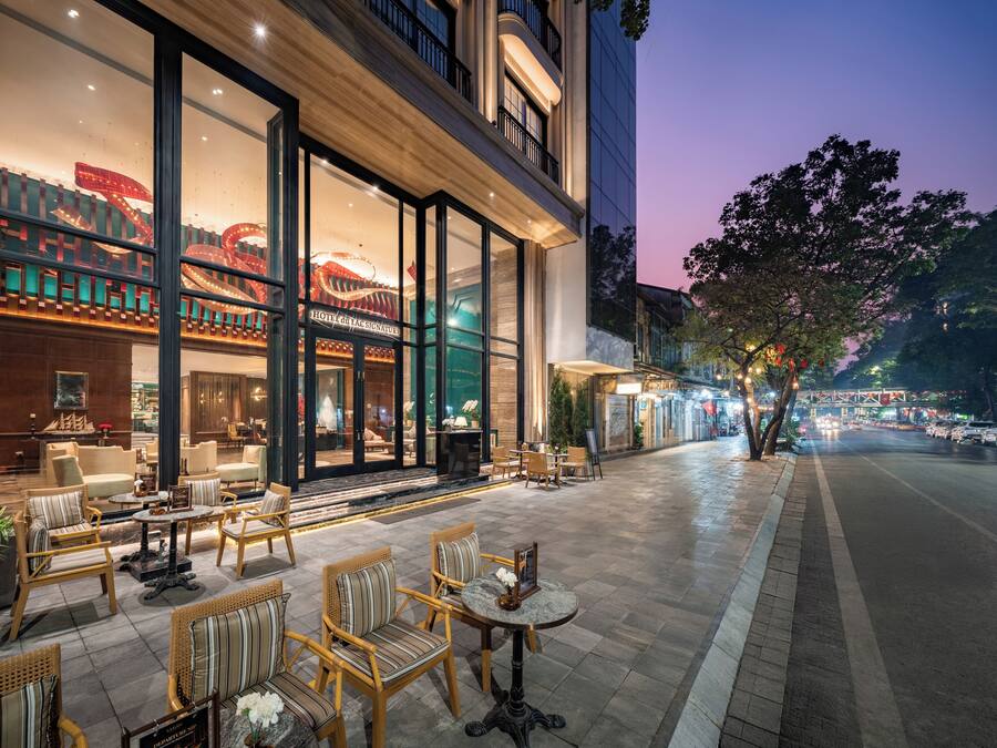Un hôtel doté d’une façade en verre, d’un espace extérieur aménagé avec des meubles en osier et offrant une vue sur la rue au crépuscule.