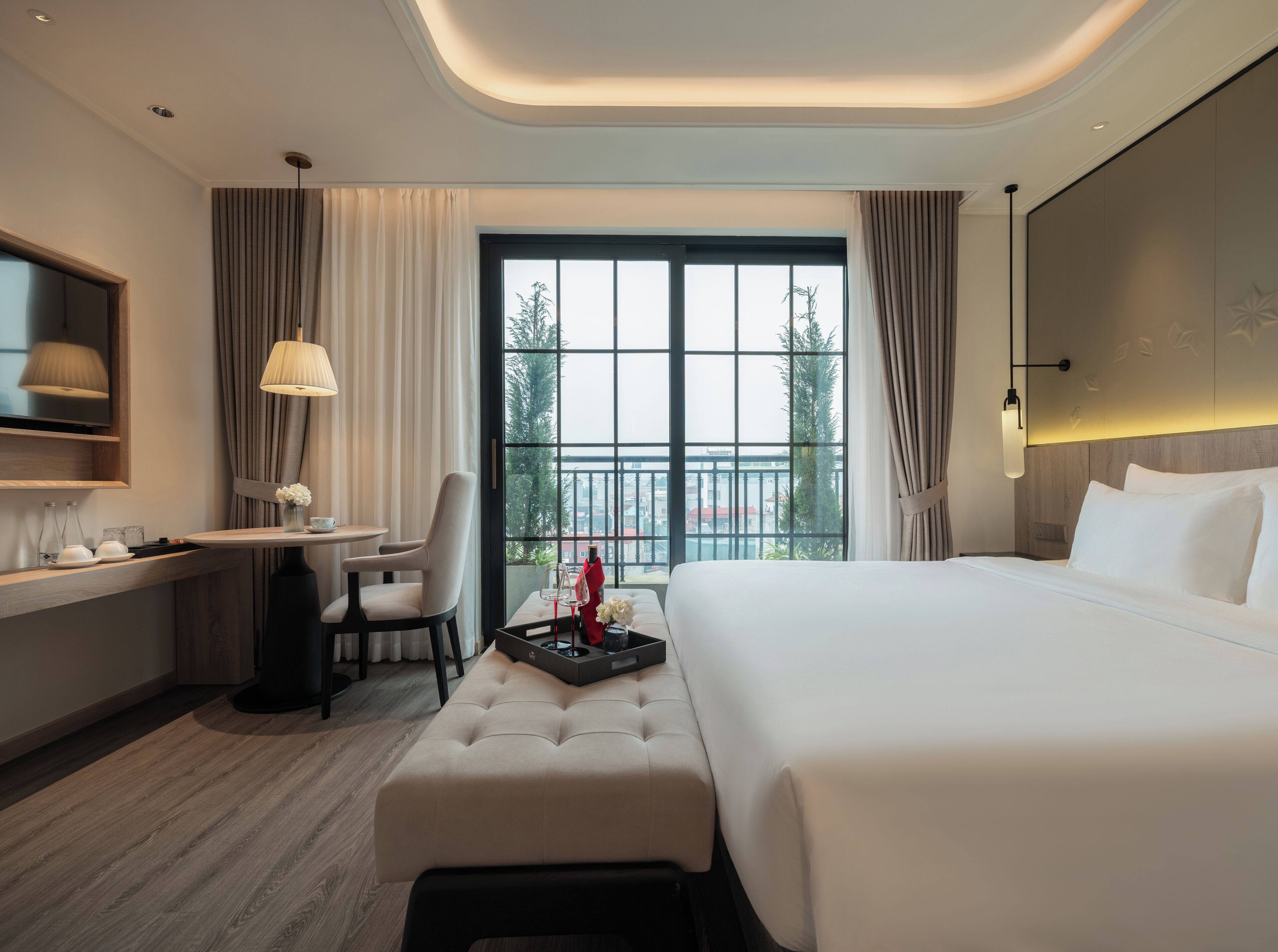 A modern hotel room with a large bed, a desk with a chair, a small table with a vase of flowers, and a large window with curtains.