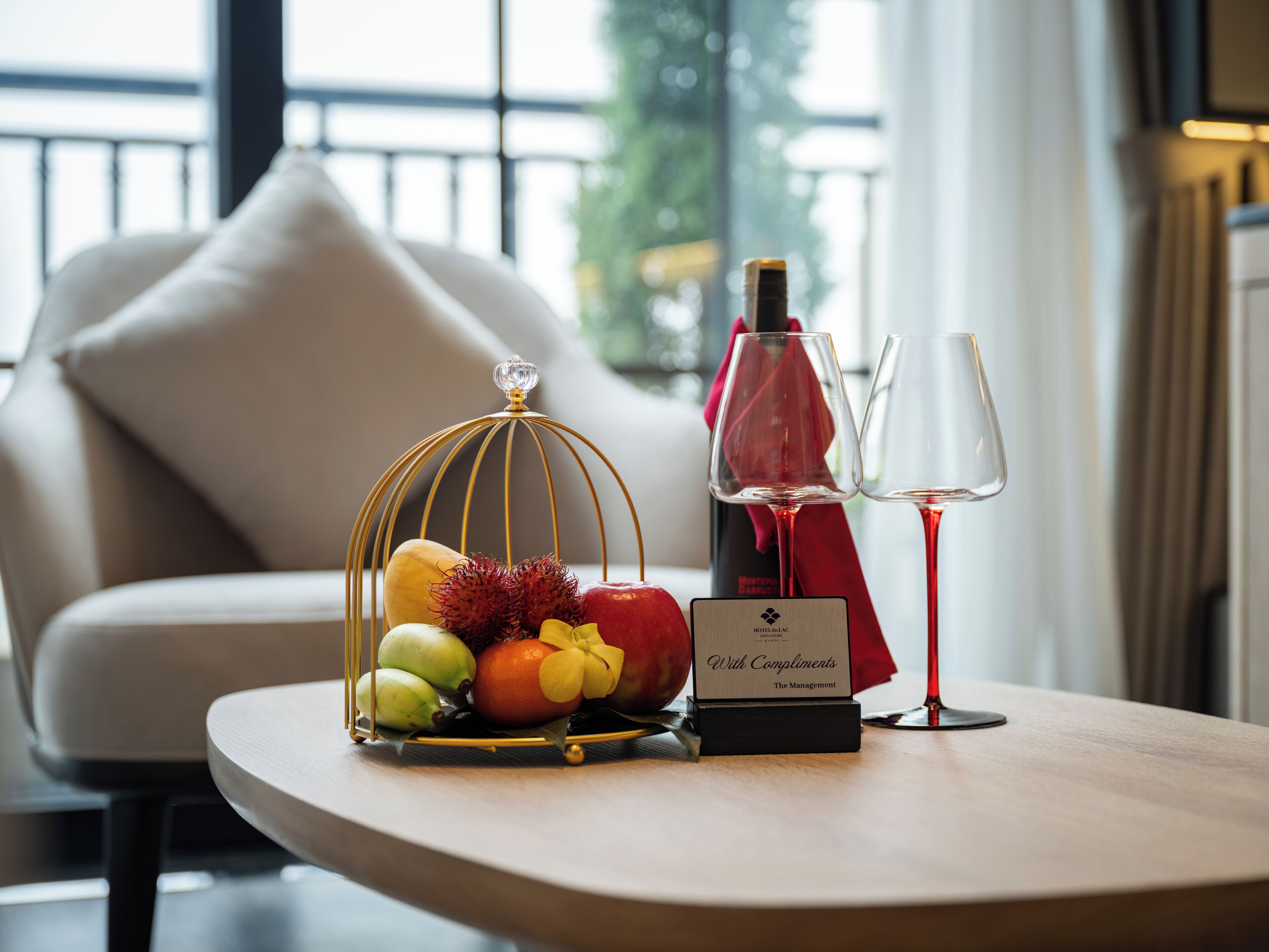 A table with a fruit arrangement, a bottle of wine, and two glasses, set against a modern interior with a sofa and a window in the background.