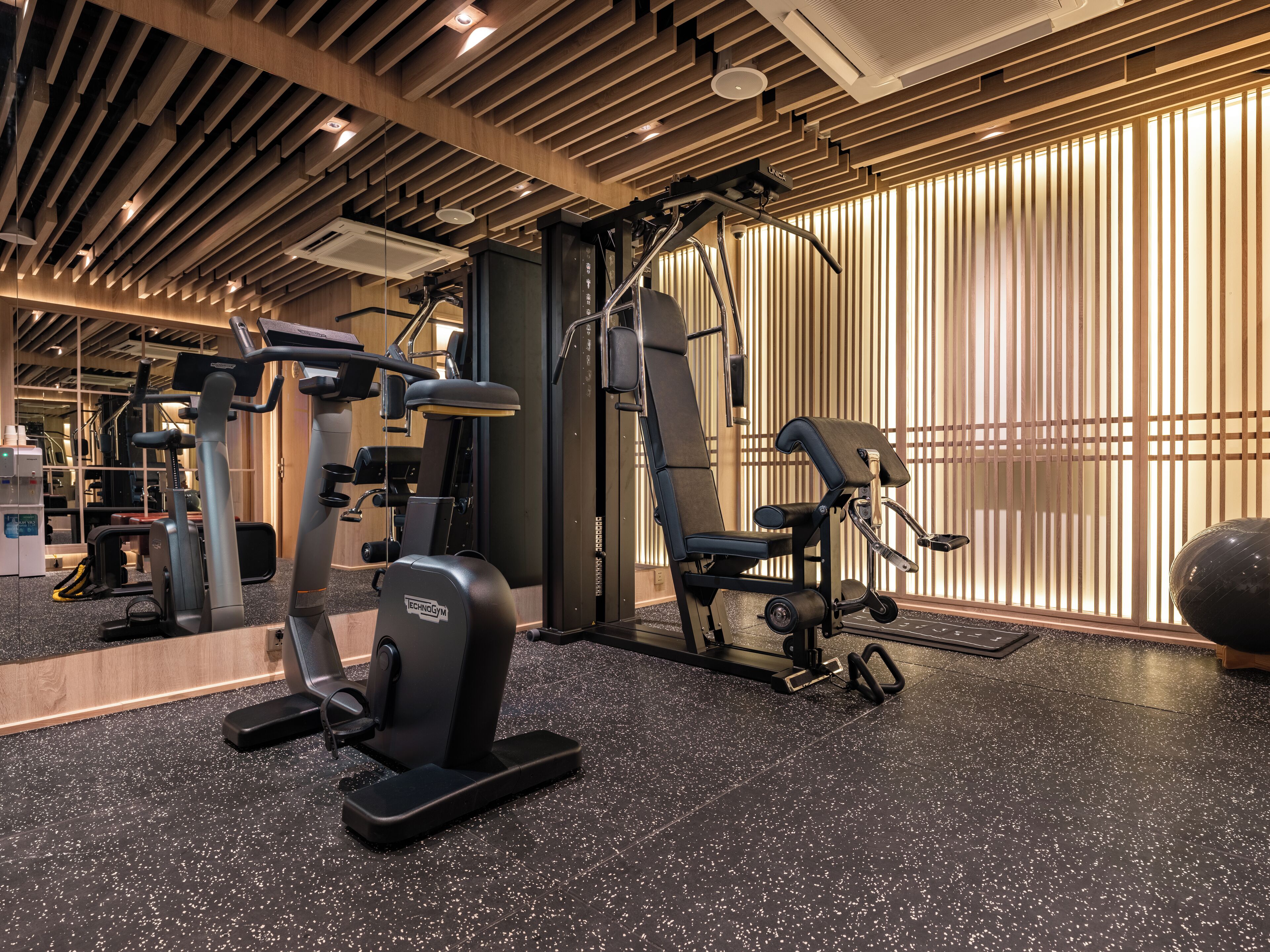 A modern gym with various exercise equipment, a speckled black floor, and wooden paneling on the walls.