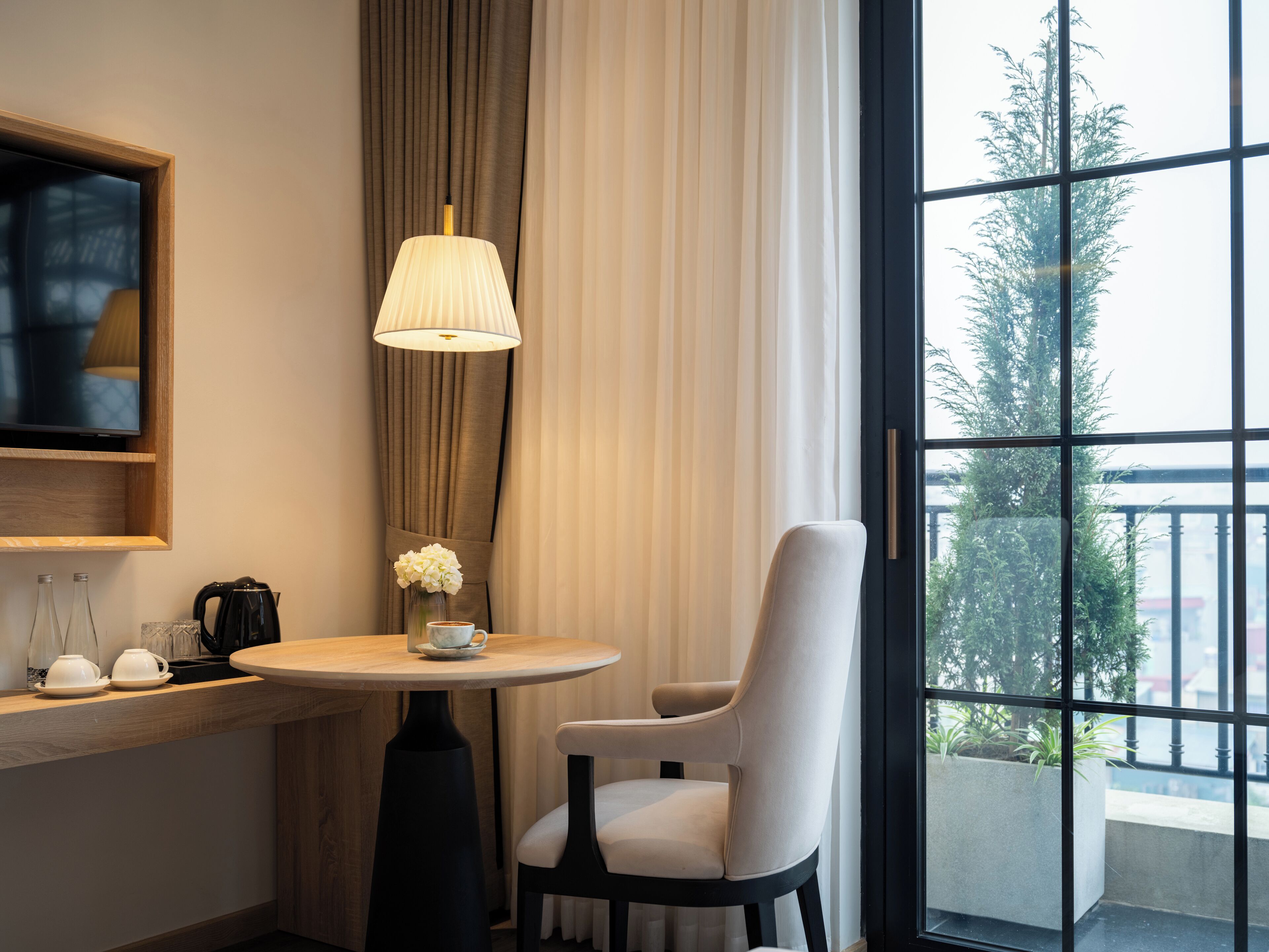 A hotel room with a round table, a chair, a lamp, a coffee maker, and a TV on the wall.