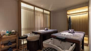 Un cuarto de spa con dos camillas de masaje, una bañera grande y un estante de madera con artículos de aseo.