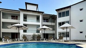 Une piscine entourée de chaises longues et de parasols, elle-même bordée d’un immeuble à plusieurs étages doté de balcons.