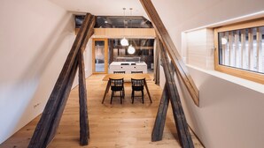 Ein Loft-Zimmer mit freiliegenden Holzbalken, Holzboden, einer Essecke mit zwei Stühlen und einem Fenster mit Jalousien.