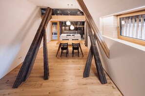 A loft-style room with exposed wooden beams, a wooden floor, a dining area with two chairs, and a window with blinds.