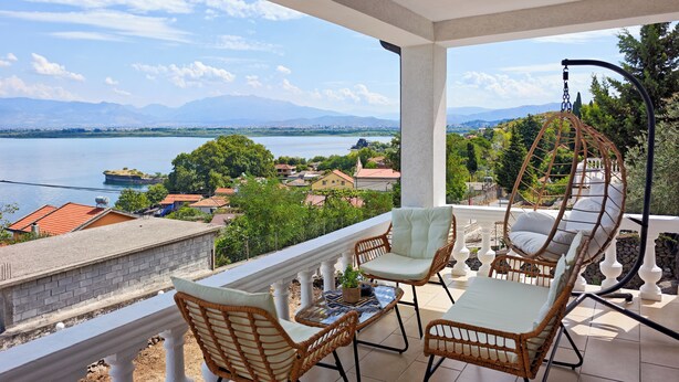 A balcony with wicker furniture, a hanging chair, and a view of a lake and distant mountains. - Villa Balani (Shiroka)