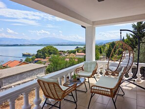 A balcony with wicker furniture, a hanging chair, and a view of a lake and distant mountains. - Villa Balani (Shiroka)