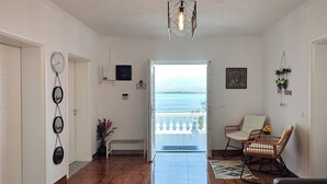 A spacious room with a tiled floor, a view of the sea through an open door, a hanging light fixture, and a chair with a cushion. - Villa Balani (Shiroka)