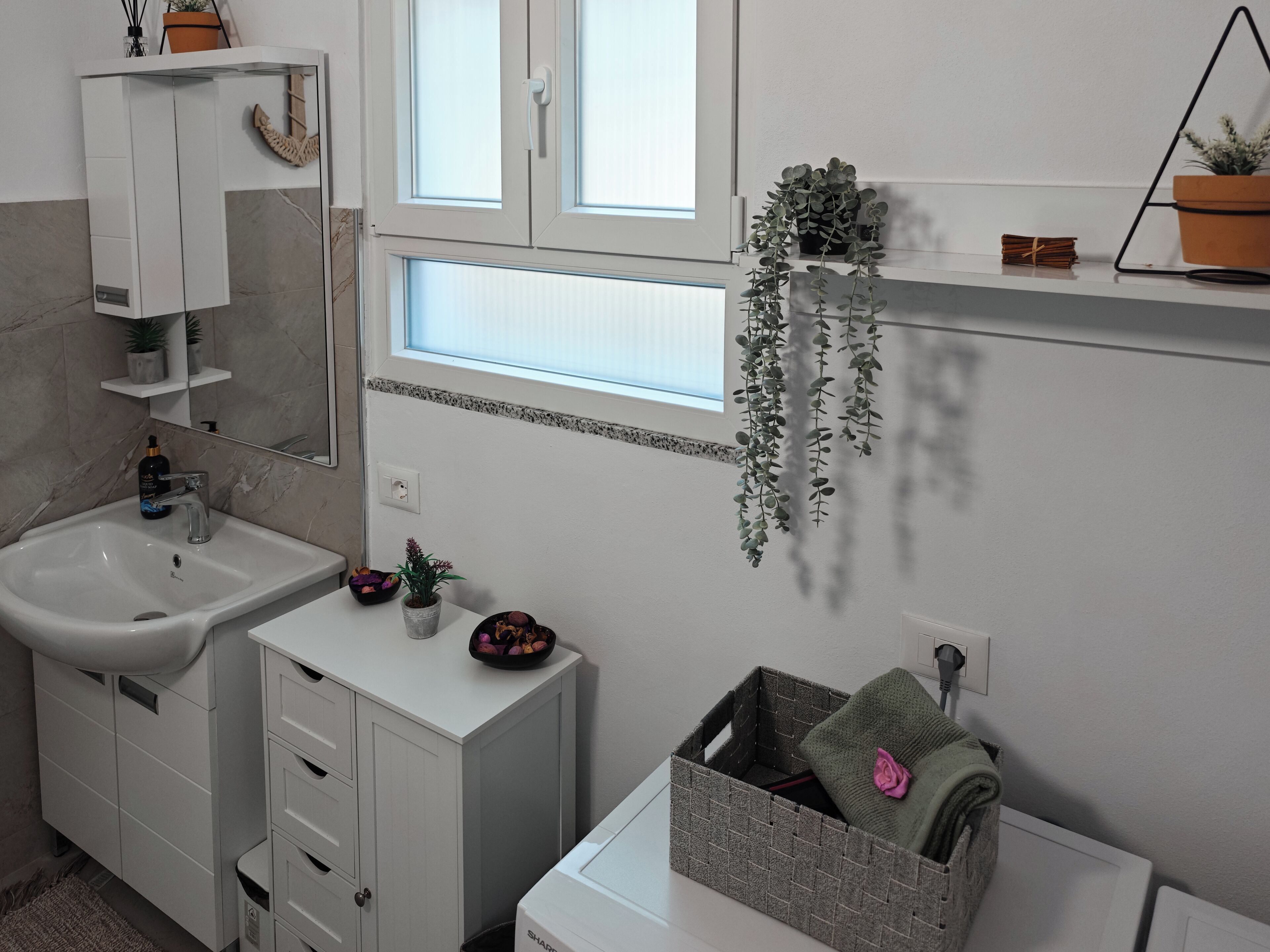 Une salle de bain moderne avec un lavabo blanc, un miroir et une fenêtre. On peut voir des plantes sur le rebord de la fenêtre et un porte-serviettes.