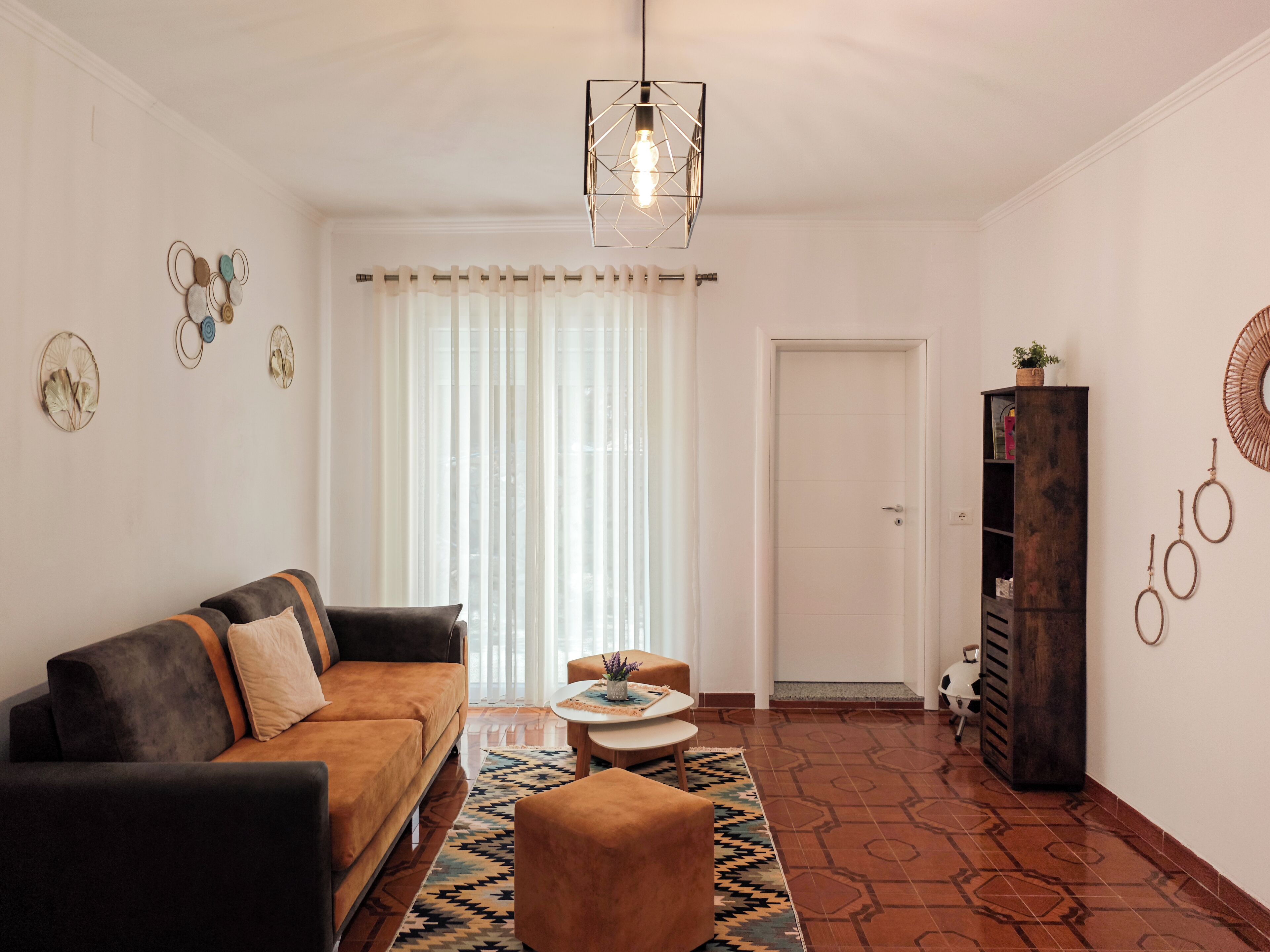 A living room with a sofa, ottoman, and a coffee table. There is a bookshelf, a door, and a window with curtains.