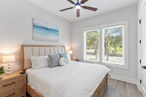 Une chambre à coucher comprenant un lit, des tables de chevet, un ventilateur de plafond, une fenêtre avec des stores et un tableau accroché au mur.