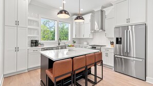 A modern kitchen with white cabinets, stainless steel appliances, a central island with bar stools, pendant lighting, and a window with a view of greenery. - 6 Ensuite Bedrooms, Pool, Close to Beach, Luxury (Santa Rosa Beach)