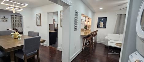 Uma cozinha moderna com piso de madeira escura, uma sala de jantar com mesa e cadeiras de madeira, e uma sala de estar com sofá branco e mesa de centro.