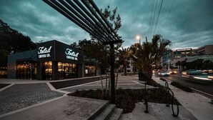 A modern coffee shop with a dark facade and illuminated signage, located in a well-lit urban area with a paved walkway and street in the background.