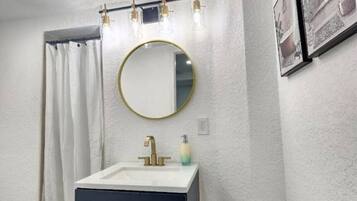 A bathroom with a white sink, dark cabinet, and a round mirror. There are light fixtures above the mirror and a framed picture on the wall.