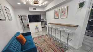 Una sala de estar moderna con un sofá azul, un tapete estampado, una zona de cocina con barra y un televisor montado en la pared.