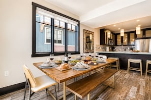 A dining area with a wooden table set for a meal, a window with blinds, and a kitchen in the background. - Tranquil Blackstone 3688 Getaway Walk to Cabriolet Lift (Park City)