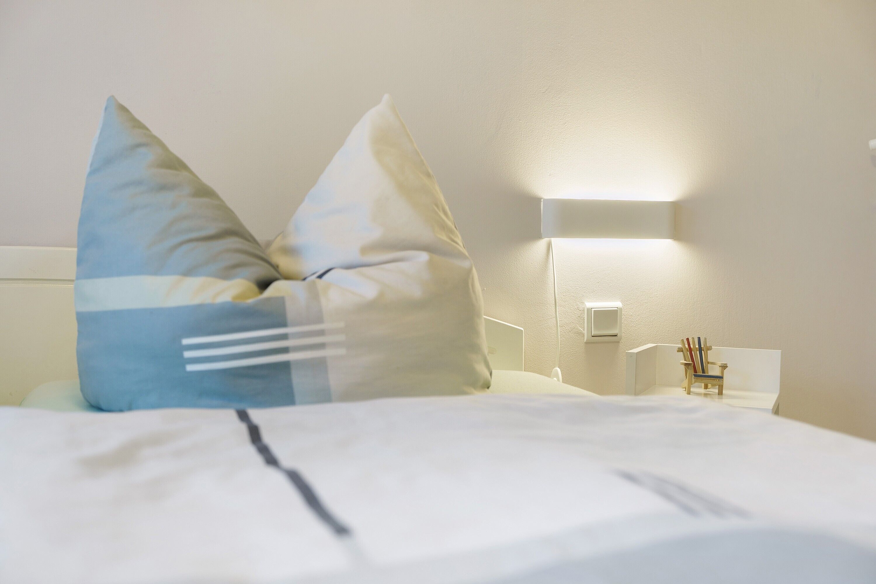 Ein ordentlich bezogenes Bett mit Kissen, ein Nachttisch mit Lampe und ein kleiner Stuhl.
