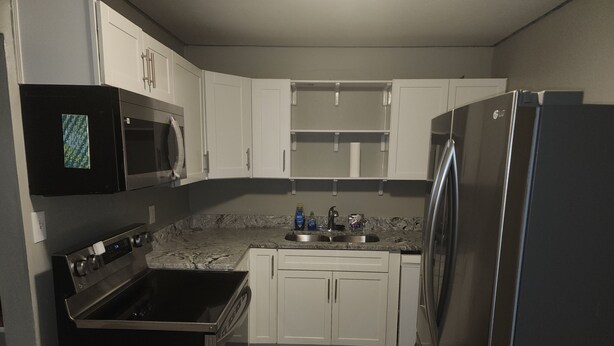 A compact kitchen with white cabinets, a black microwave, a stainless steel refrigerator, and a granite countertop.