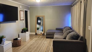 A living room with a flat-screen TV, a sofa, a small white cabinet, a mirror, and a door leading to another room.