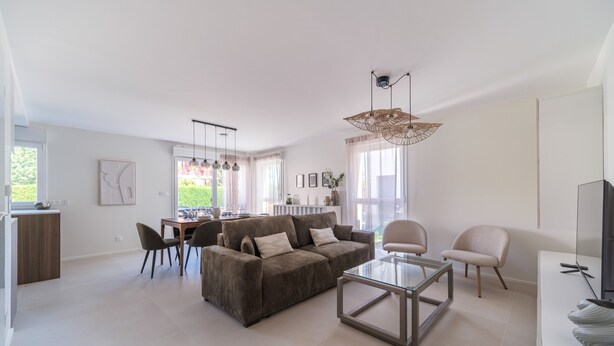A modern living room with a sofa, coffee table, and chairs. There is a dining area with a table and chairs. The room has large windows with curtains.