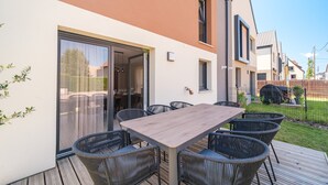 A modern outdoor patio with a wooden table and wicker chairs, a sliding glass door leading to a covered area, and a view of neighboring houses.