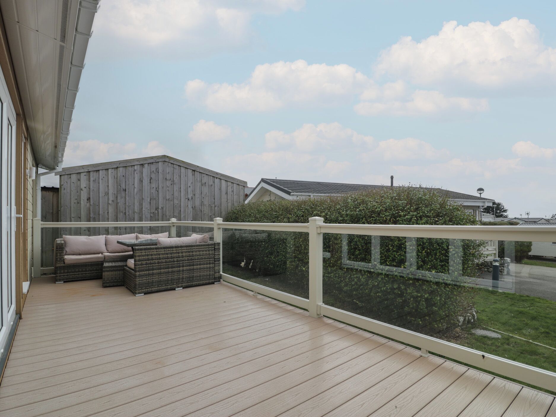 Een houten terras met een witte reling, een rieten bank met kussens, en uitzicht op een woonwijk met huizen en een haag.