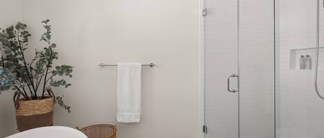 Un baño con ducha de vidrio, una bañera blanca, una cesta de mimbre y una toalla blanca en un perchero.