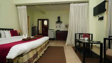 A hotel room with a bed, a small table with a vase, a TV mounted on the wall, and a kitchenette in the background.