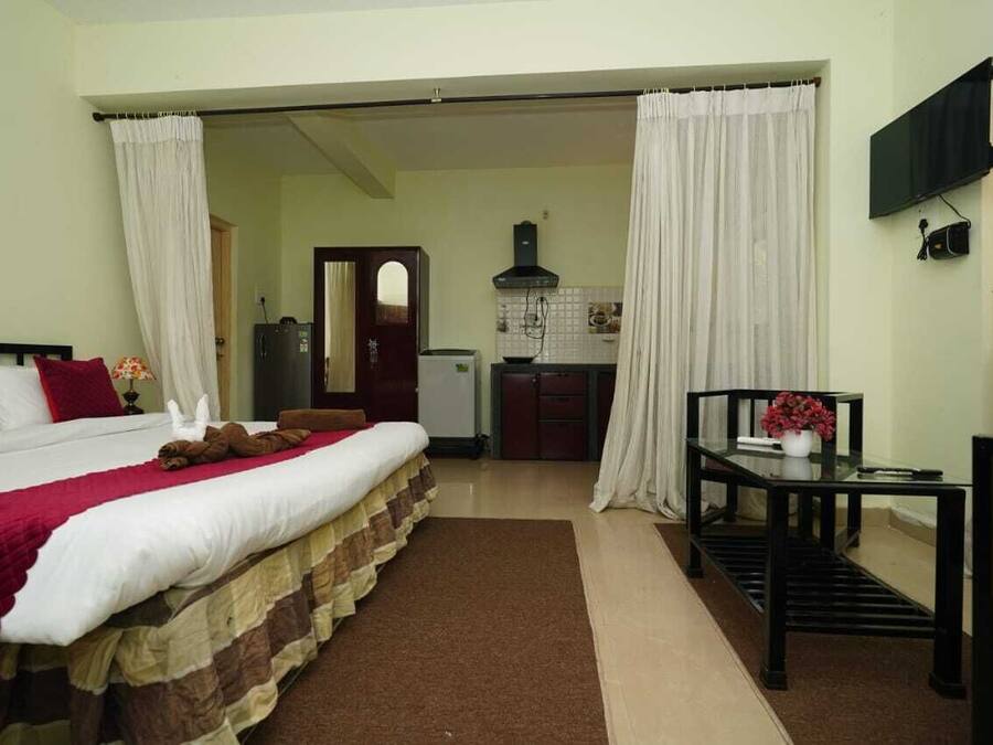 A hotel room with a bed, a small table with a vase, a TV mounted on the wall, and a kitchenette in the background.