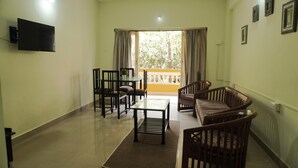 A living room with a TV mounted on the wall, a glass coffee table, and two sofas. There is a balcony with curtains and a view of greenery. - Four Seasons GuestHouse (Utorda)