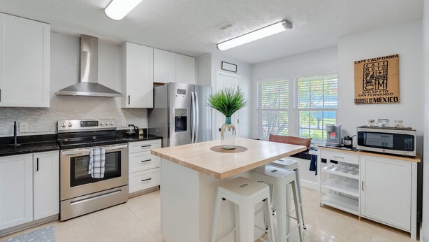 A modern kitchen with white cabinetry, stainless steel appliances, a central island with stools, and a window with blinds. - The Dive Locker- 5 min to the Beach Sleeps 10 (Panama City Beach)