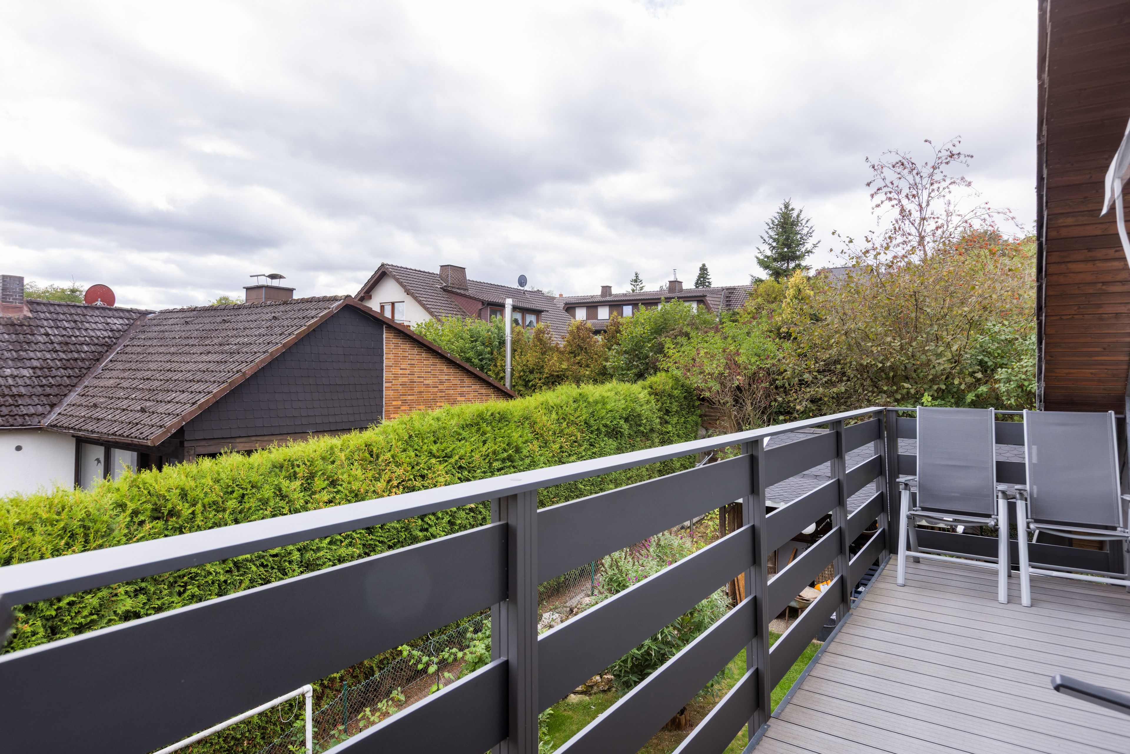 Eine Holzterrasse mit Metallgeländer, mit Blick auf ein Wohngebiet mit Häusern und einer Hecke.