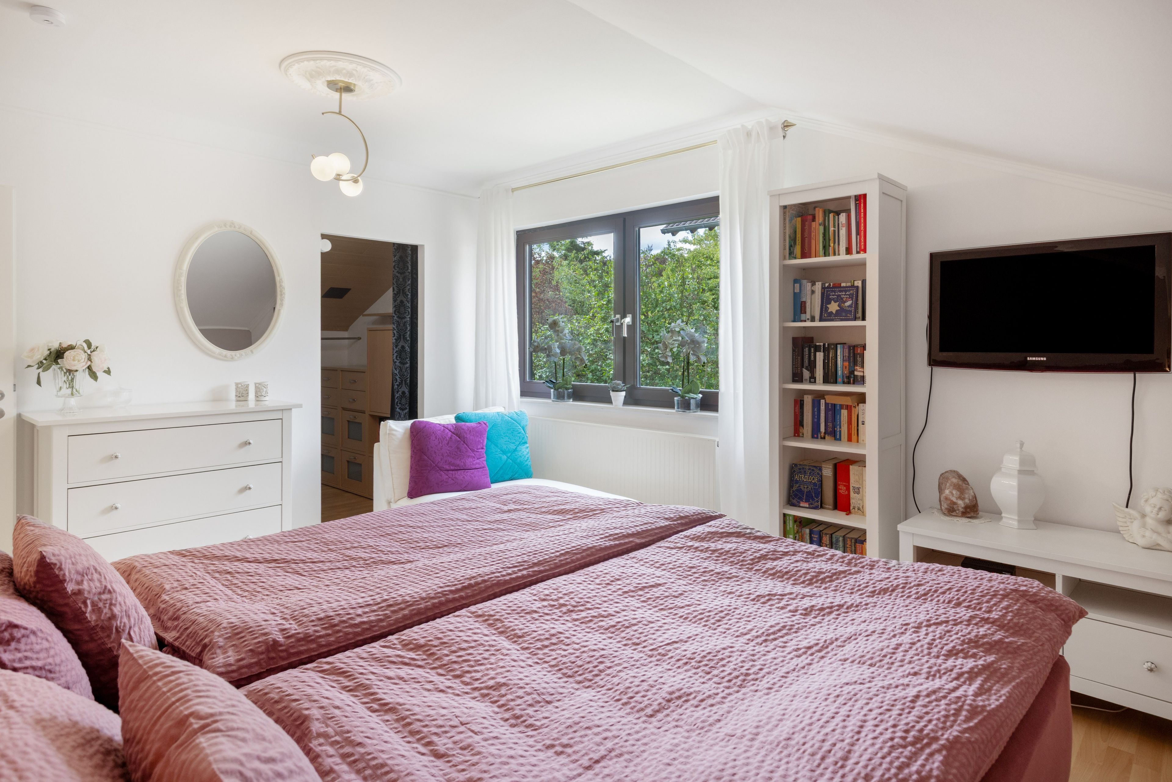 Ein Schlafzimmer mit zwei Betten, einem Bücherregal, einem Fernseher und einem Fenster mit Blick auf Bäume.